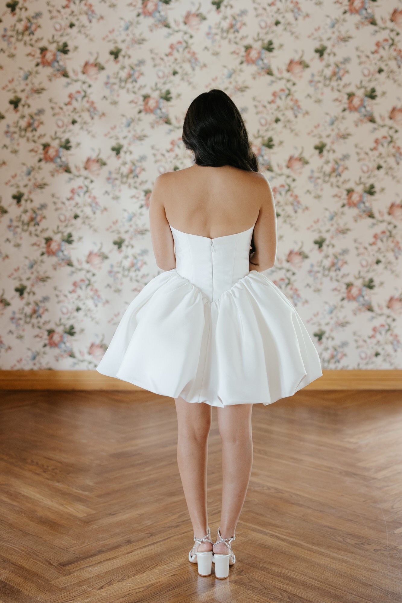 Woman in a white strapless dress standing in a room with floral wallpaper.