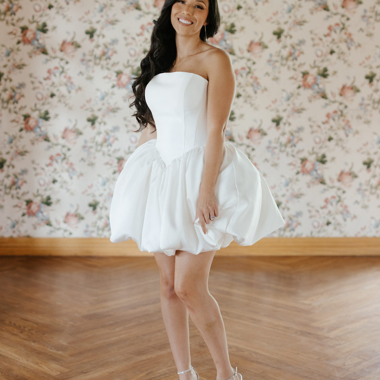 Woman in a white strapless dress standing in a room with floral wallpaper.