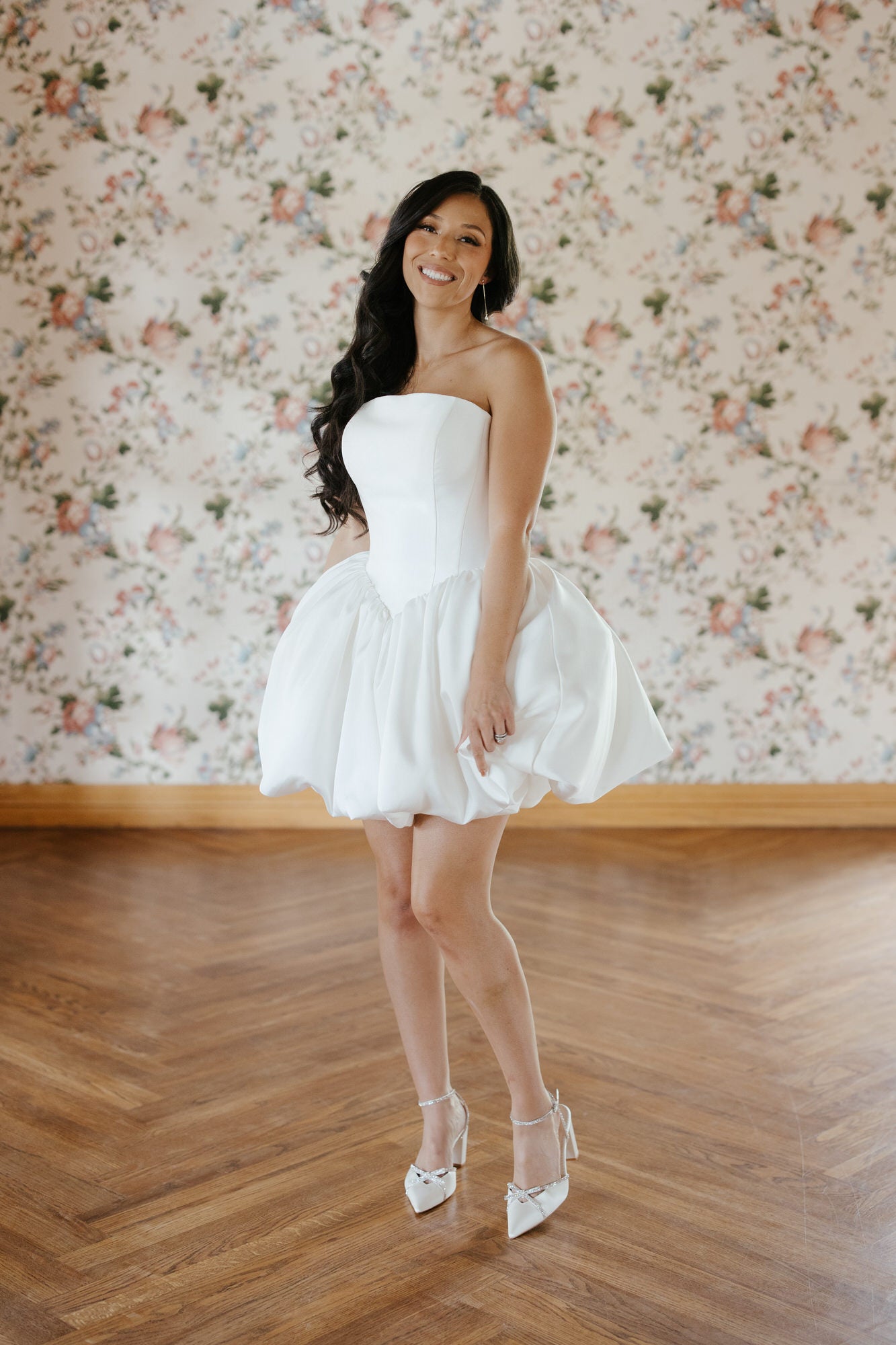 Woman in a white strapless dress standing in a room with floral wallpaper.