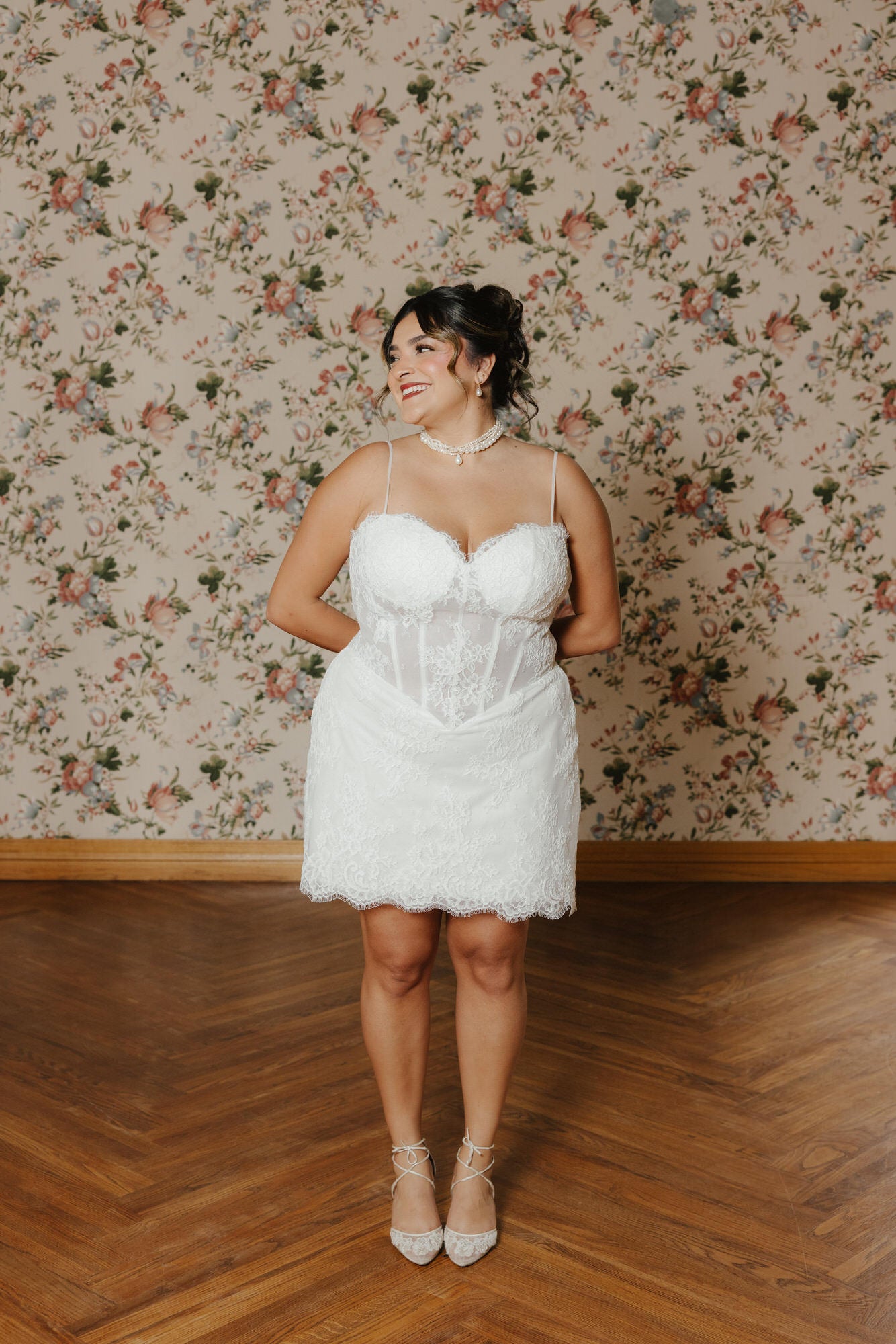 Woman in a white lace dress standing in front of floral wallpaper