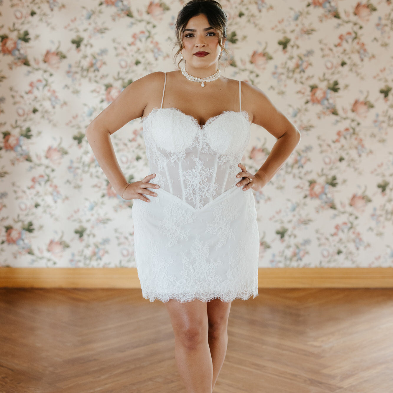 Woman wearing a white lace dress standing in front of floral wallpaper.