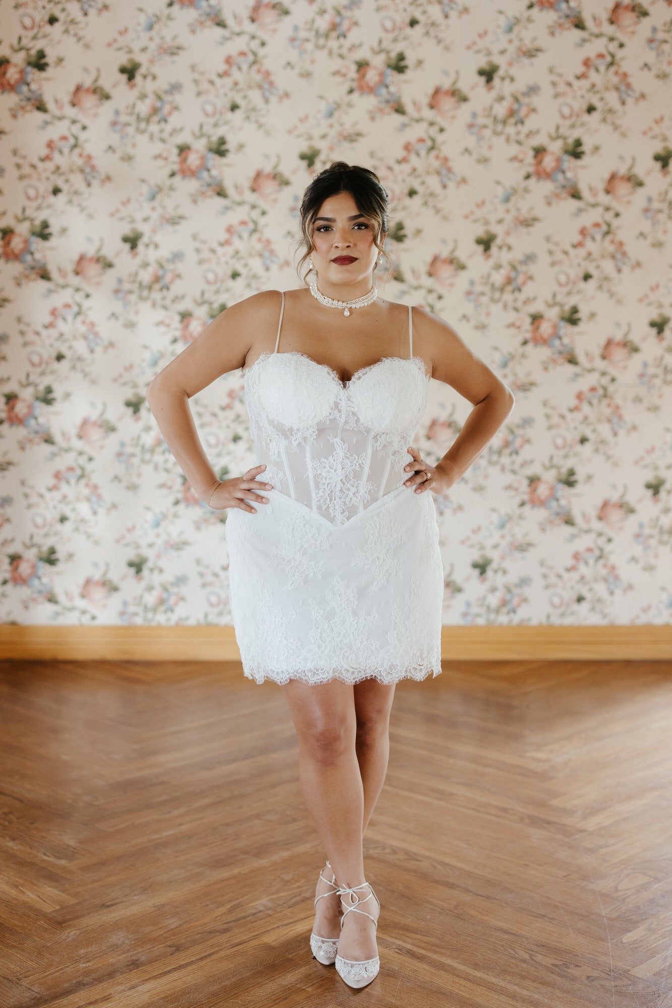 Woman wearing a white lace dress standing in front of floral wallpaper.