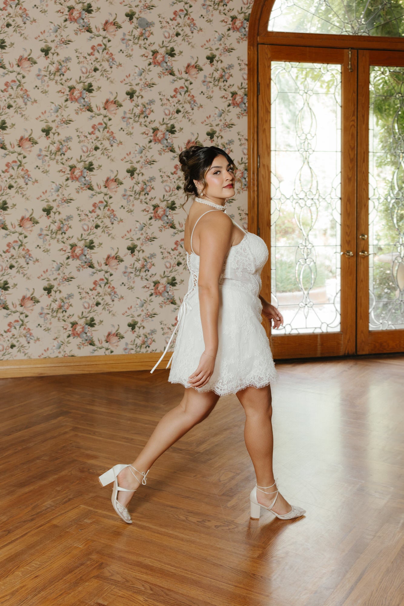 Woman in a white lace dress standing in a room with floral wallpaper and a large window.
