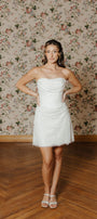 Woman in a white strapless dress standing against a floral wallpapered wall.