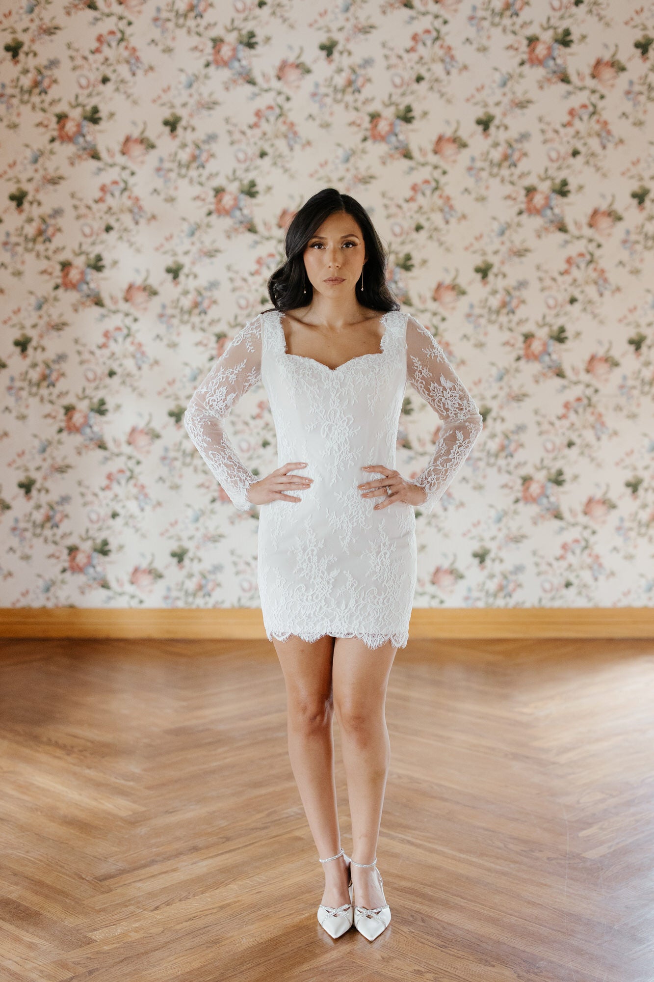 Woman in a white lace dress standing in front of floral wallpaper