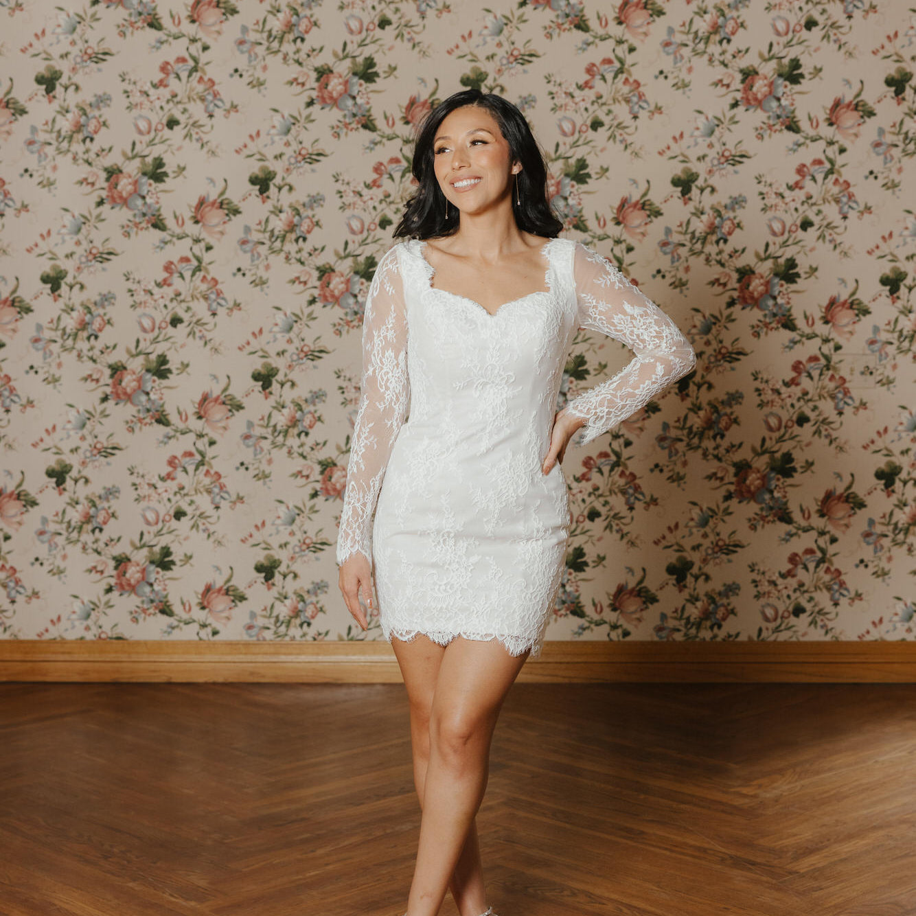 Woman in a white lace dress standing in a room with floral wallpaper.