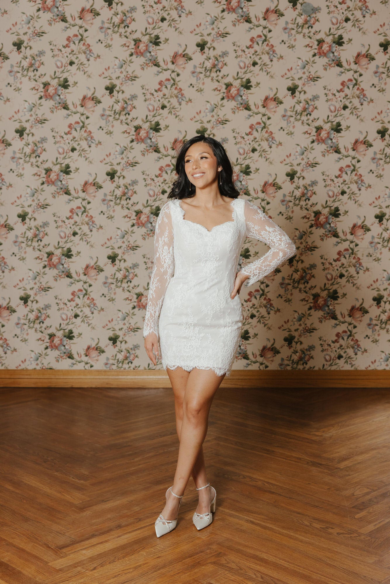 Woman in a white lace dress standing in a room with floral wallpaper.