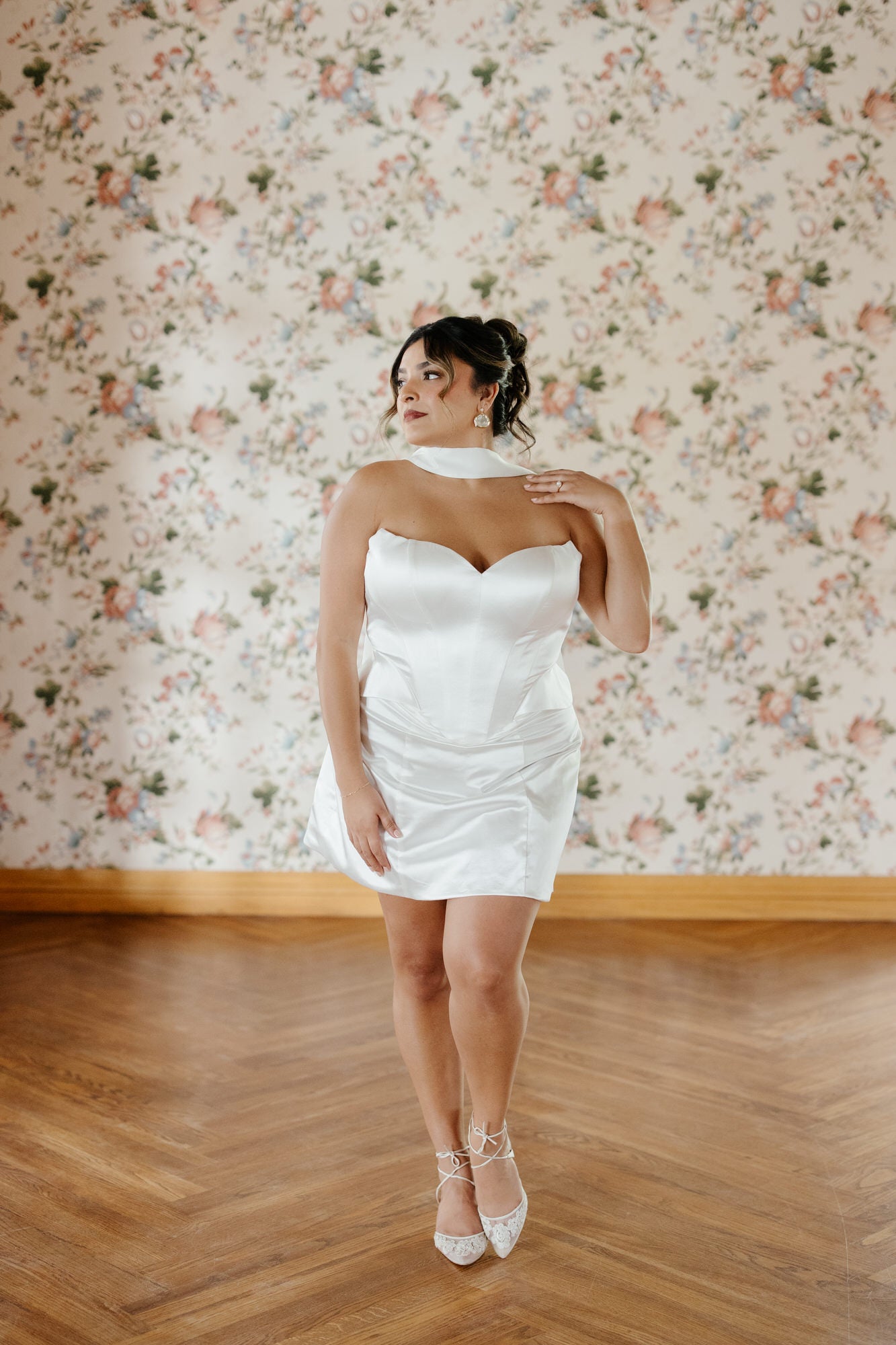 Woman in a white dress standing against a floral wallpapered wall.