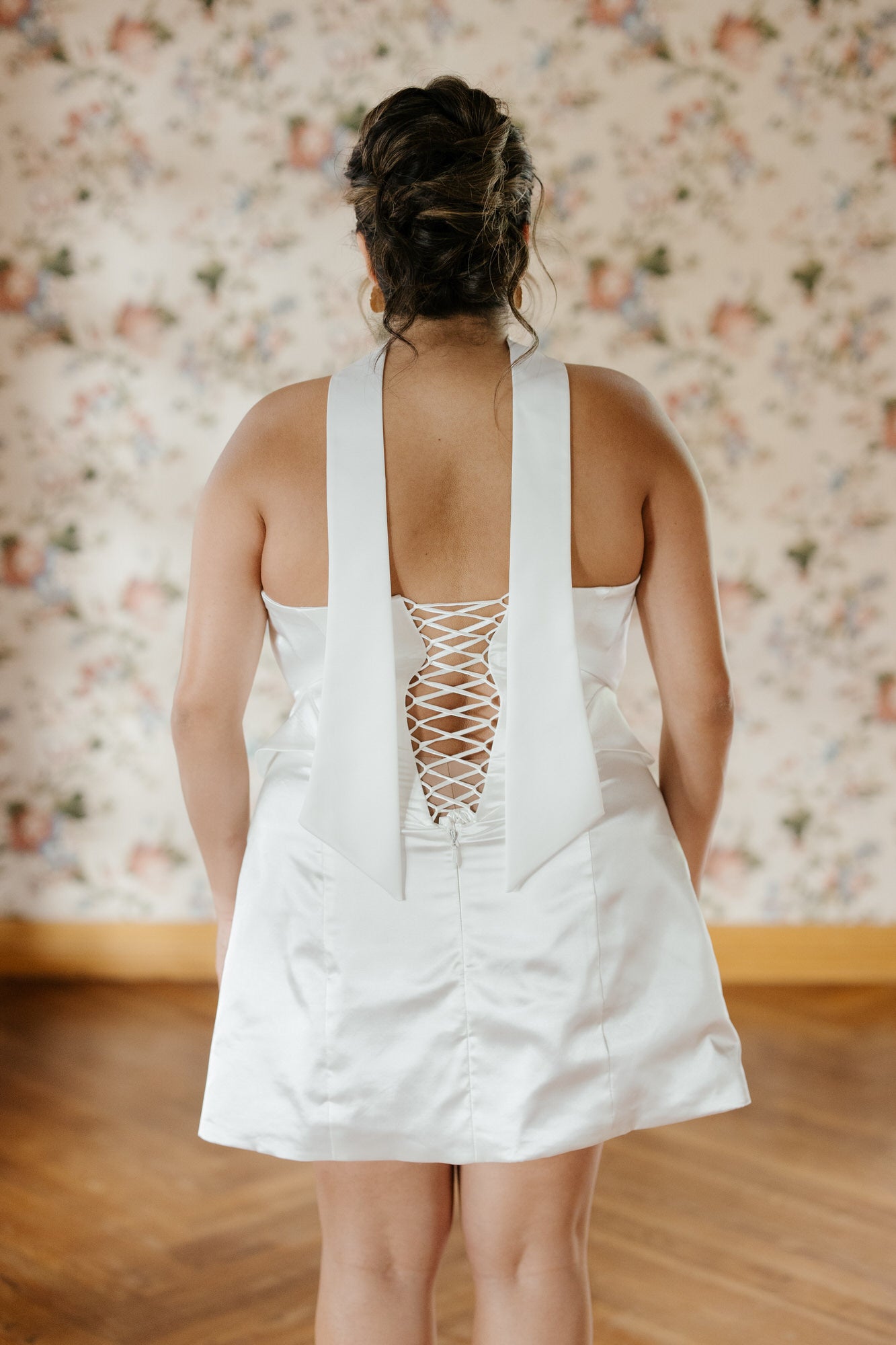Person wearing a white dress with a lace-up back in a room with floral wallpaper.