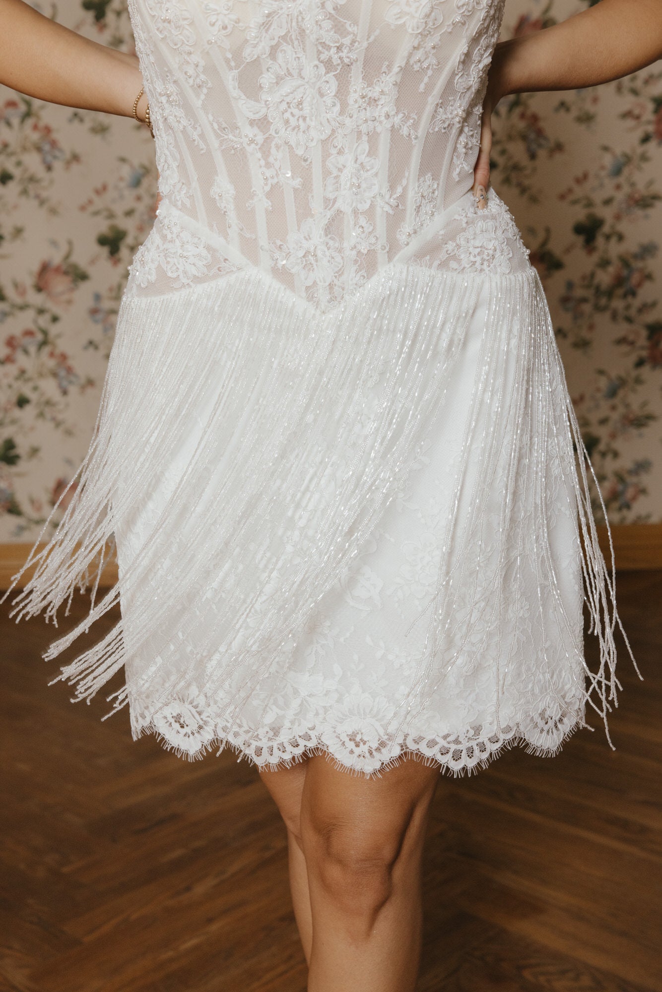 White lace dress with fringe details against a floral wallpaper background