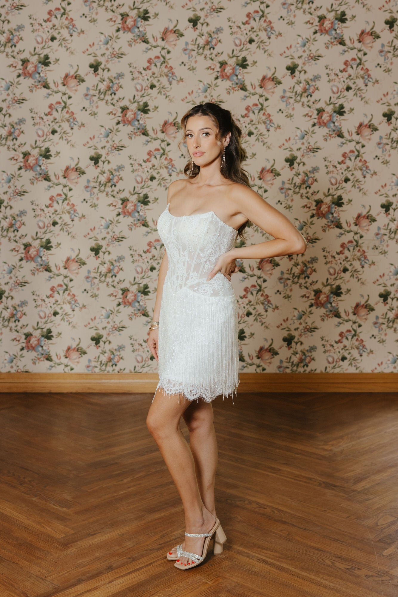 Woman in a white strapless dress standing against a floral wallpapered wall.