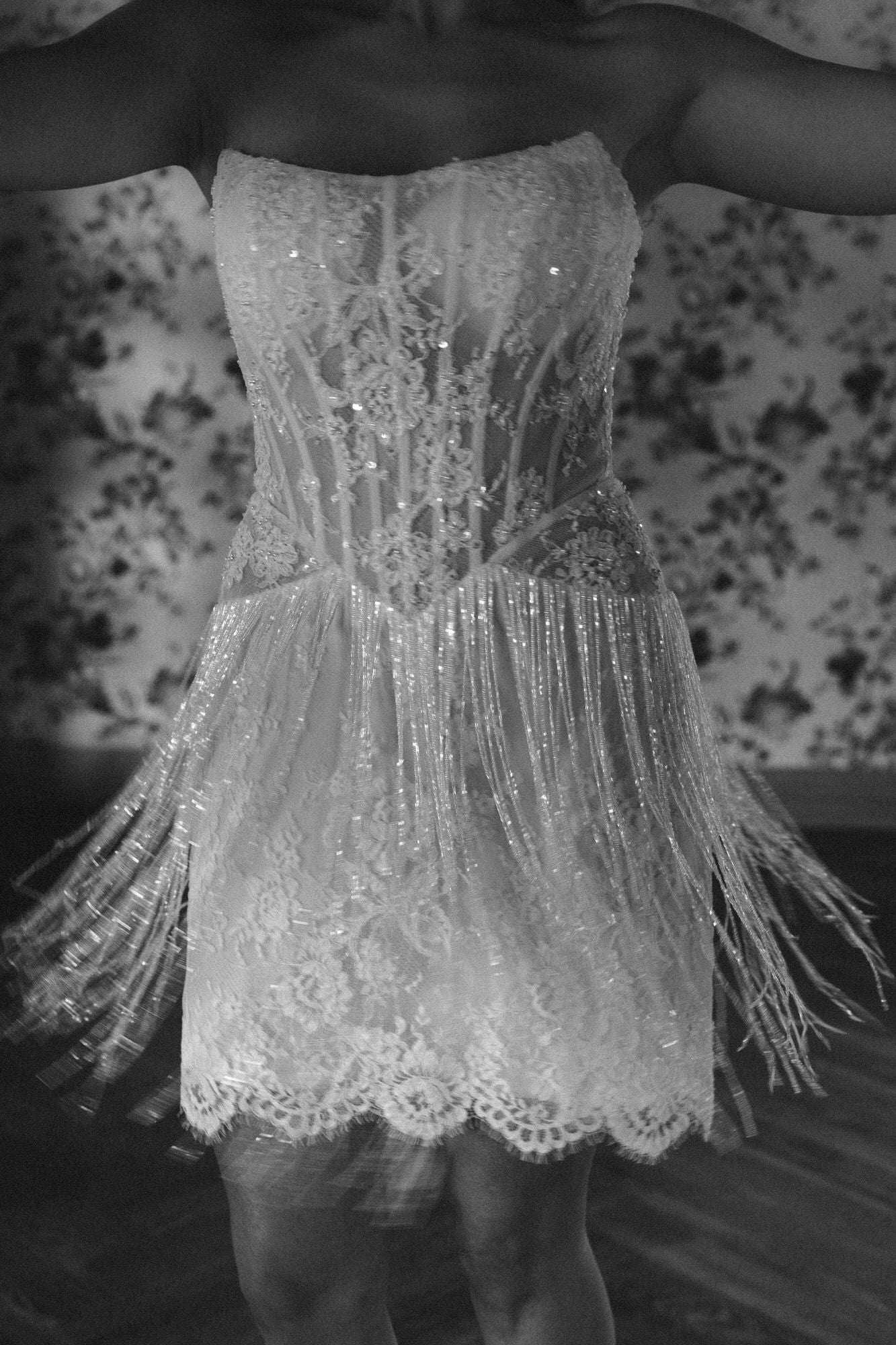 Person wearing a sparkling, strapless dress with fringe details against a floral-patterned wall.