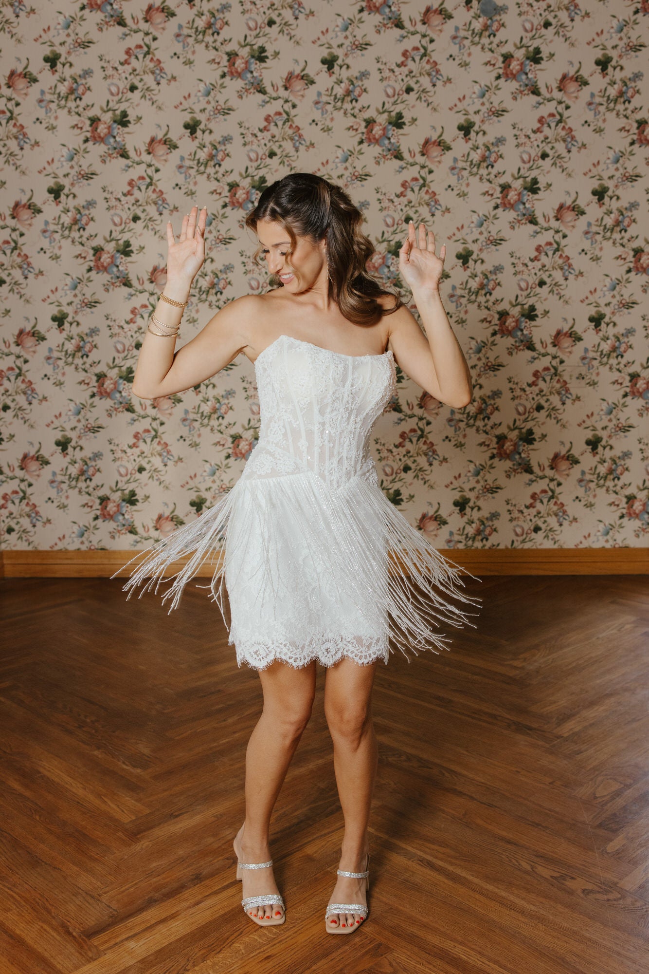 Woman in a white lace dress with fringe standing in a room with floral wallpaper.