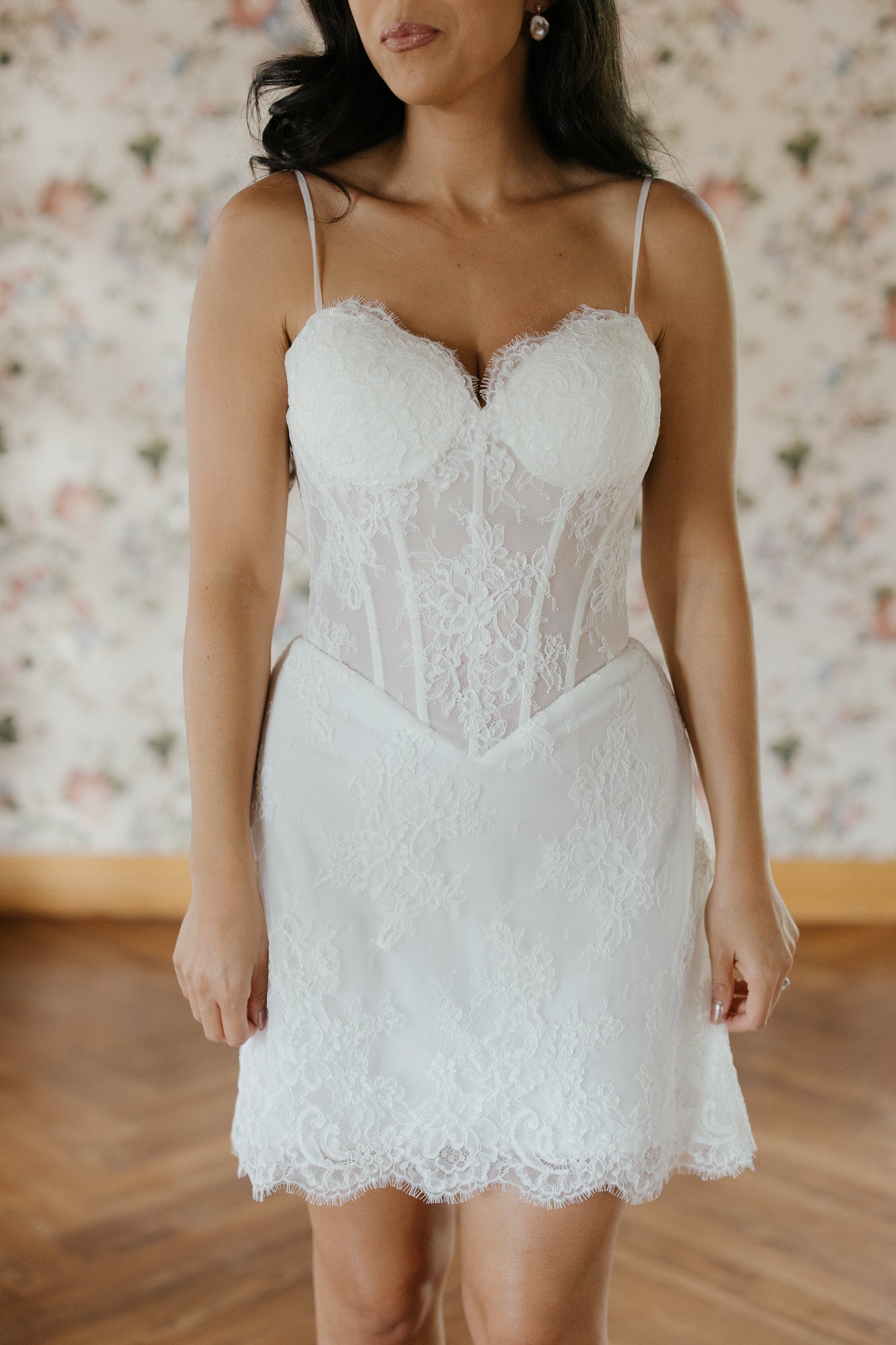 Woman wearing a white lace dress against a floral wall.