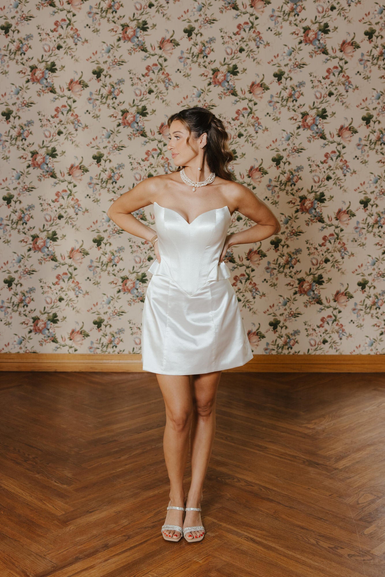 Woman in a white strapless dress standing against a floral wallpapered wall.