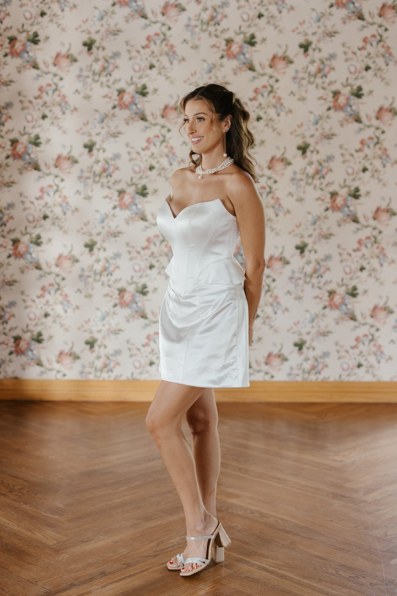 Woman in a white strapless dress standing against a floral wallpapered wall.
